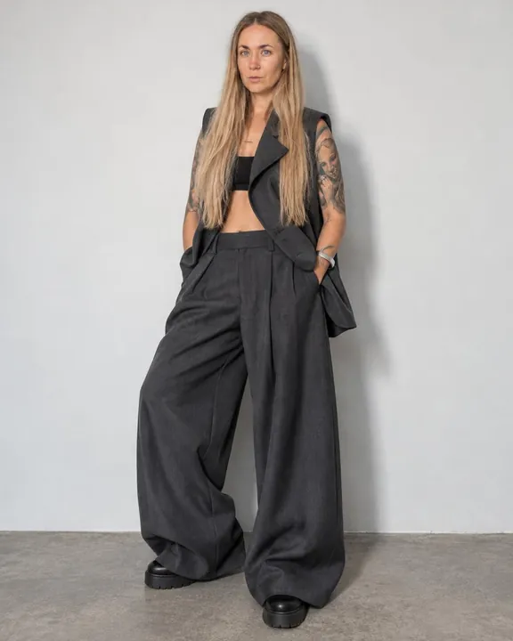 Full-length woman with long blond hair against a plain wall, sleeveless charcoal jacket, very wide trousers and chunky boots