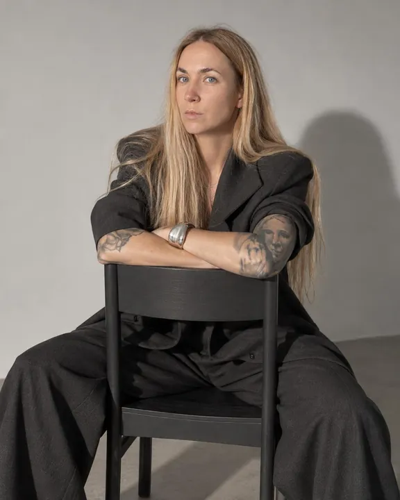 A woman with long blond hair sits backwards on a black chair, arms folded on the backrest, a chunky silver cuff