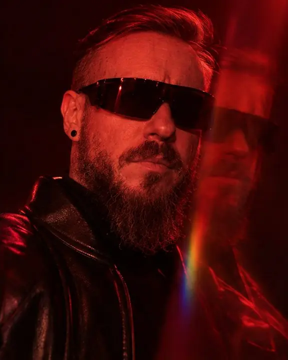 A bearded man in shield sunglasses lit deep red, a faint prism copy of his face overlapping his shoulder