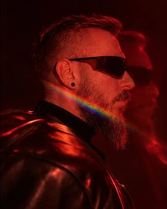 Profile of a bearded man in red light, his doubled shoulder line and a thin rainbow sliver crossing the jaw