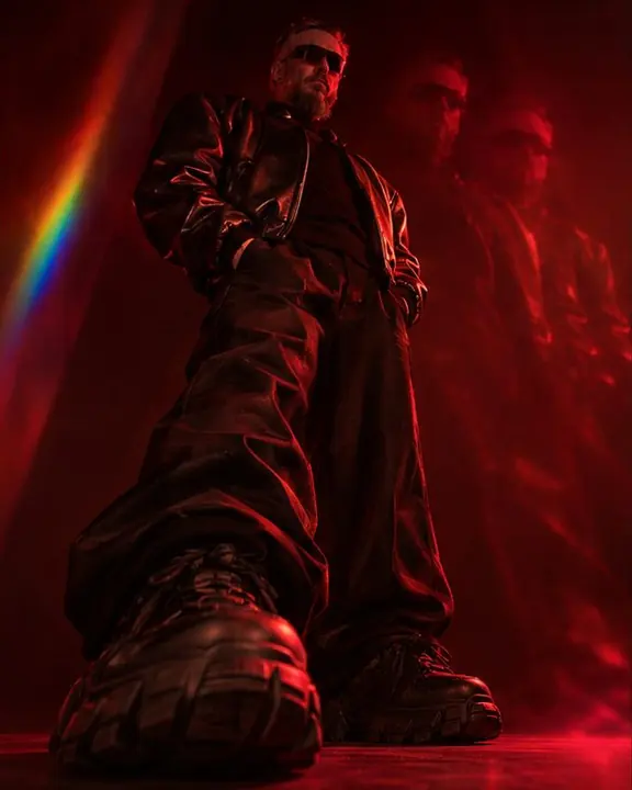 Low angle on chunky sneakers and pooled black jeans, a man's huge padded shoulders and red copies towering above
