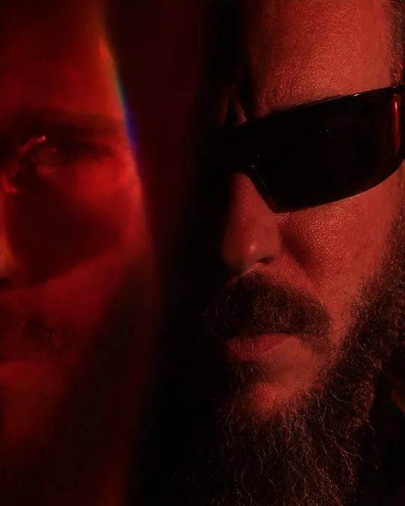 Close-up of a bearded man's face half lost in black, a red prism echo of one eye floating beside it