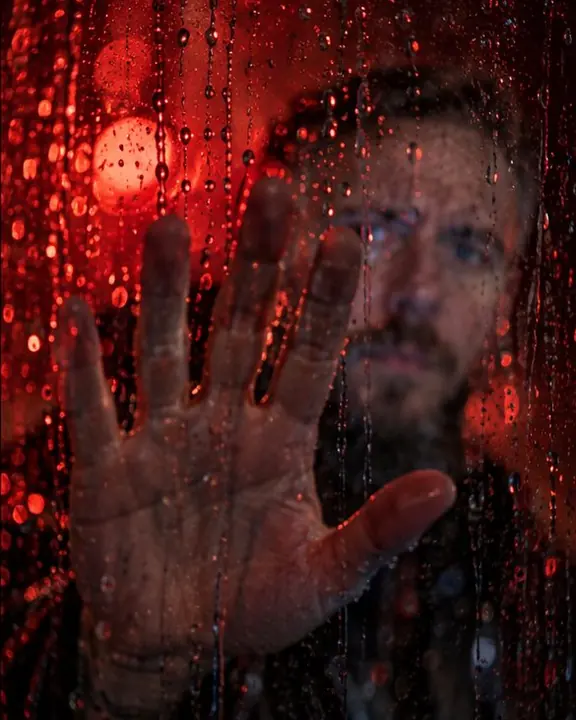 A man's hand pressed against the far side of a wet window at night, red-lit raindrops gathered around it