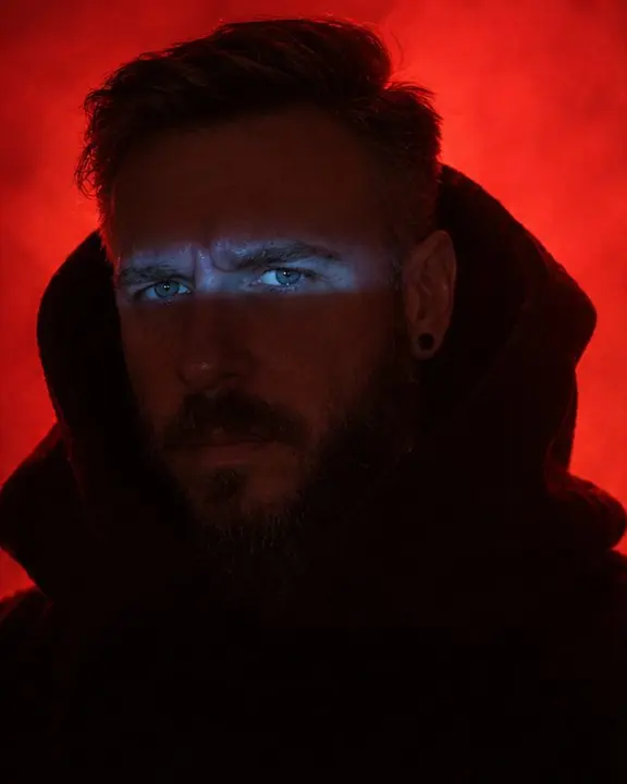 Head and shoulders of a man in a funnel hood, a narrow blue slit of light falling across his eyes