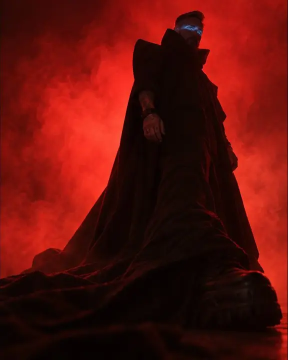 Low angle on a man's floor-length black train spreading across the floor, red smoke rising around him