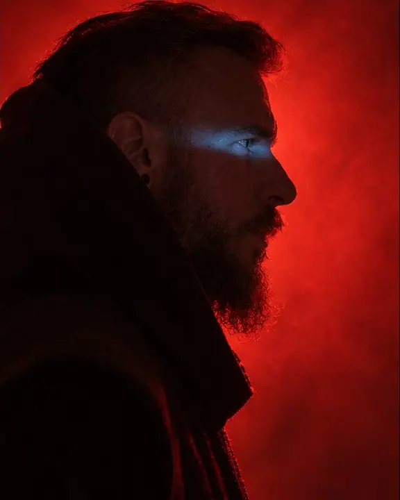 Profile of a bearded man, hood edge and beard outlined in red backlight, his eye catching a blue line