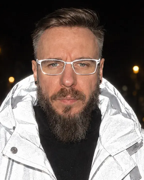 Head and shoulders of a bearded man lit by flash, a glowing collar around his face and glasses rims flaring