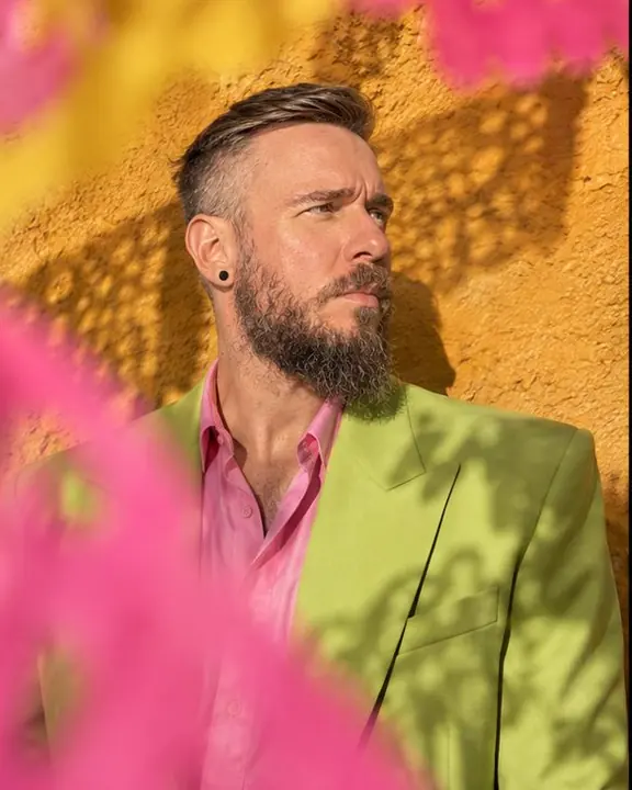 A bearded man in an acid-green suit stands against an ochre wall, seen through blurred pink and yellow paper flags