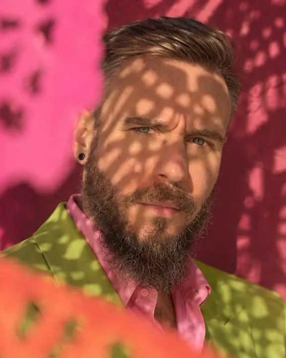 Patterned shadows from cut-paper flags fall across a bearded man's face in hard Mexico City sunlight