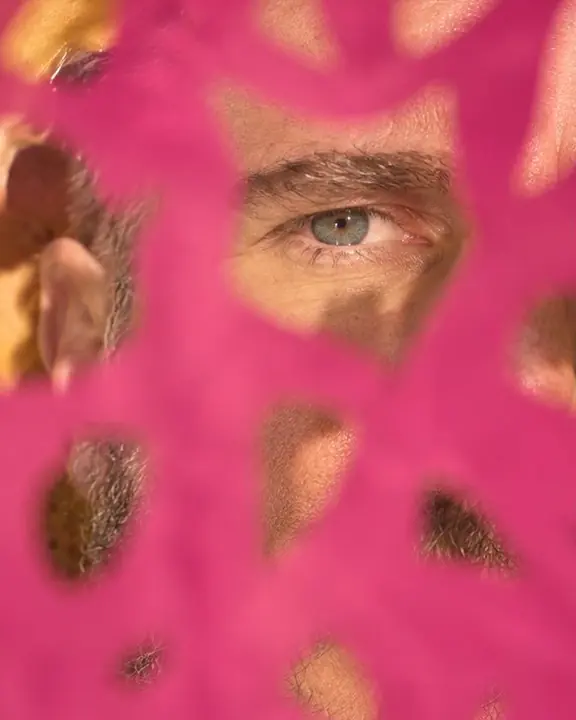 Close-up of one eye of a bearded man seen through a cut shape in a sheet of colored paper