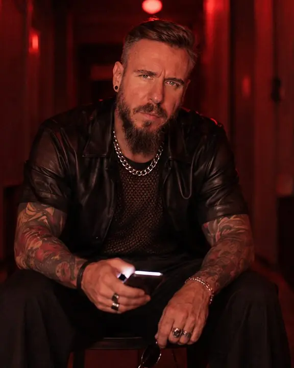 A man sits on a low stool in a red corridor, elbows on knees, a white phone screen lighting his fingers