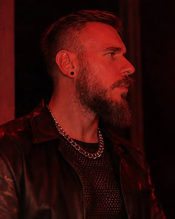 Profile of a bearded man against a black doorway, red light tracing his face, silver chains glinting at the neck