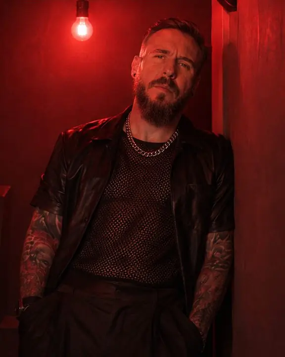 A man in black mesh and leather leans on a wall under a bare red bulb, his face lit from above
