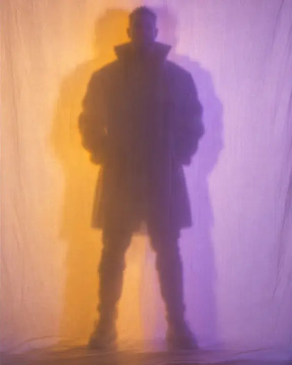 The soft shadow of a man's full figure on a glowing white scrim, doubled in amber and violet