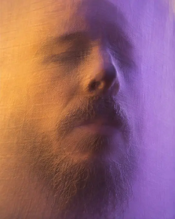 A bearded face pressed into backlit fabric, the stretched relief of nose, lips and beard showing in the cloth