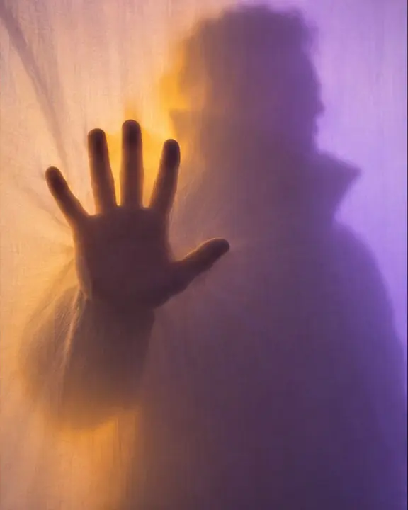A man's hand pressed flat and sharp against a backlit scrim, his body behind it a soft giant amber shadow