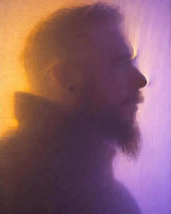 A man's profile shadow turning sharp on white fabric as his cheek touches the scrim, amber and violet glow