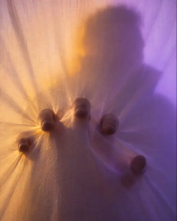 Fingertips pressing into backlit white fabric, soft folds radiating outward in amber and violet light