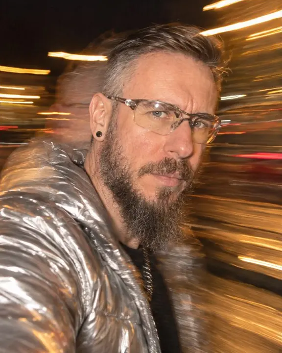 A bearded man in a silver puffer spins toward the camera, face frozen sharp, amber light trails sweeping right