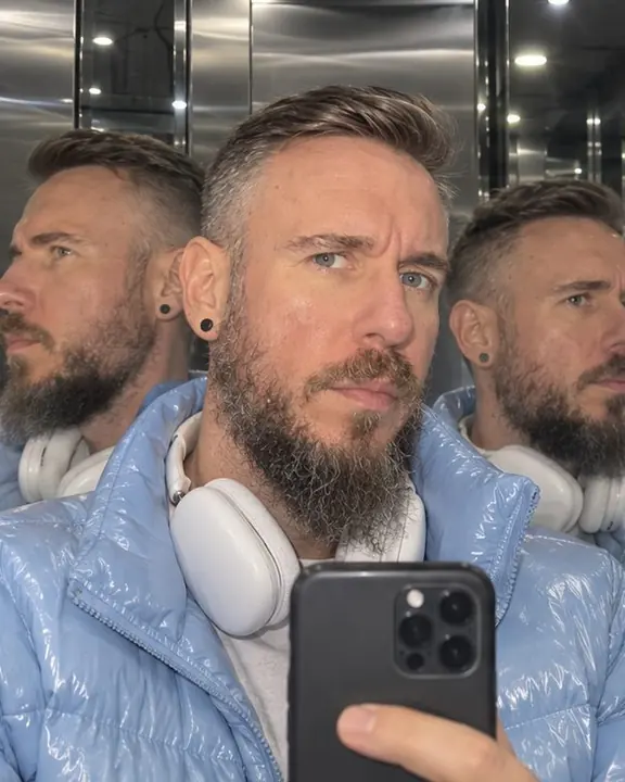 Close-up of a bearded man lowering his phone, his face reflected three times at different angles in elevator mirrors