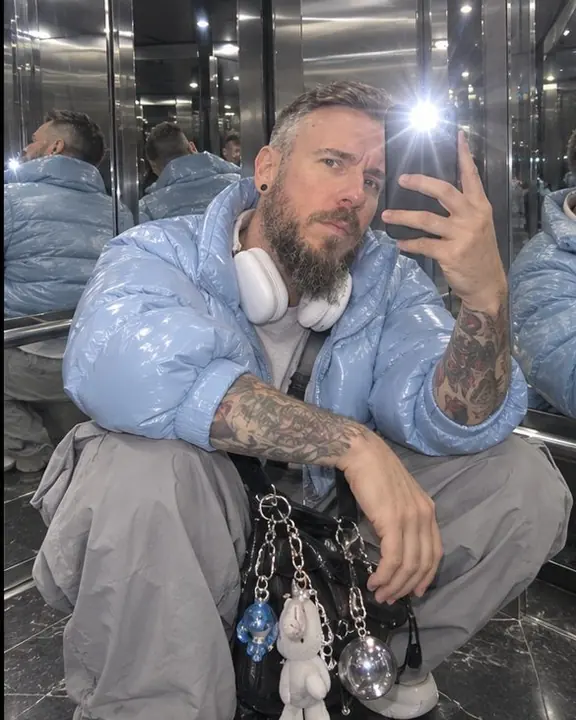 A man crouches in a mirrored elevator corner, white headphones and a charm-covered bag close to the ultra-wide lens