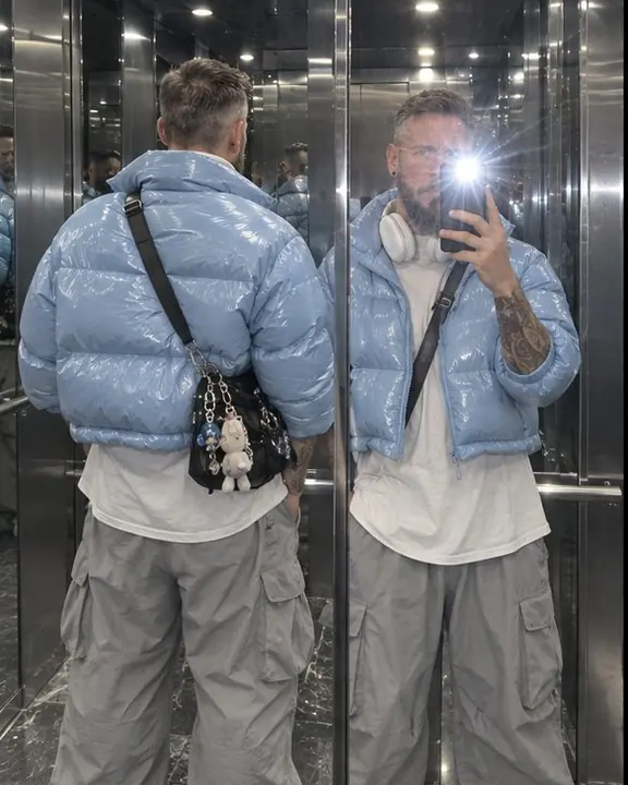 A man's back in one elevator mirror and his front in another, cool white ceiling light and silver walls