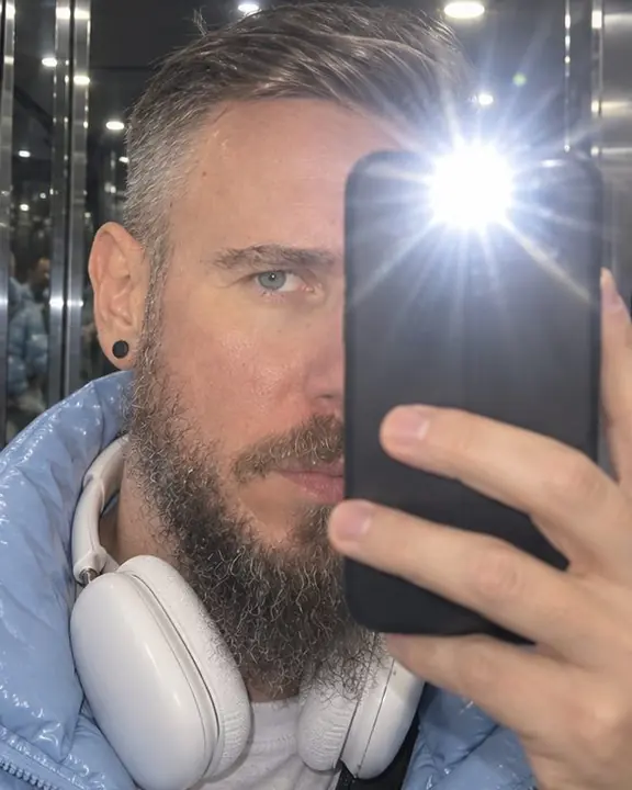 Phone flash bloom covering half of a bearded man's face in an elevator mirror, one eye still visible