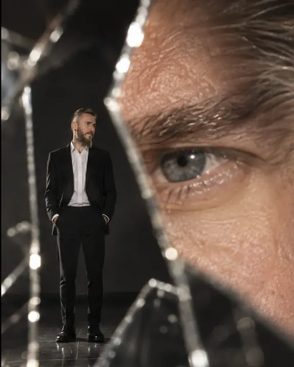 A tiny full-length man in a black jacket seen directly, beside a giant reflected eye filling a large shard