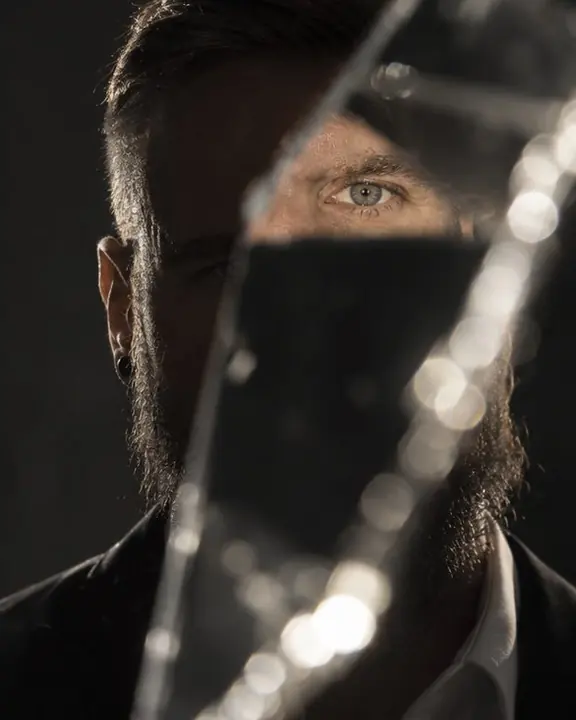 A bearded man's face mostly hidden behind a dark shard, one reflected eye looking straight at the lens