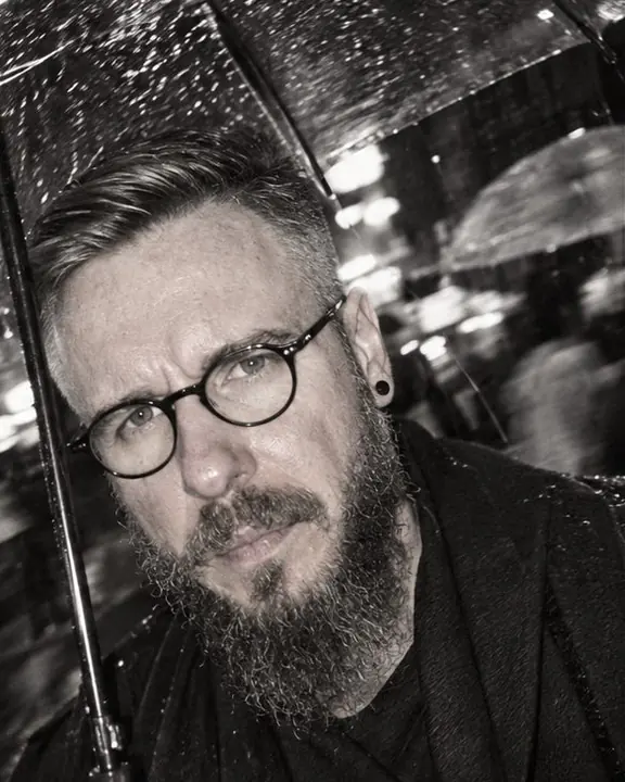 Tilted close-up of a bearded man lit by hard flash, raindrops frozen midair, blurred umbrellas behind, black and white
