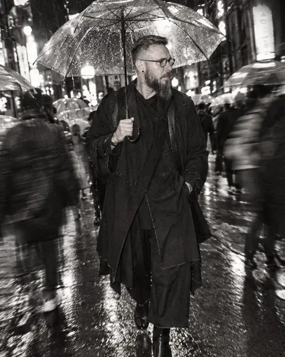 A man in a black wrap coat stands under a clear umbrella in a Tokyo alley as a rainy crowd smears past him