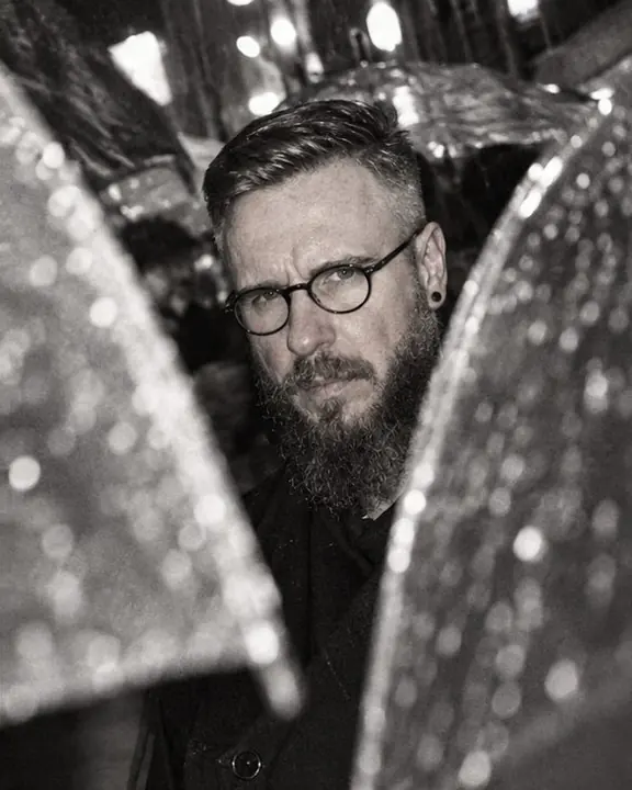 A bearded man's face in hard flash seen through a gap between two umbrellas, grainy black-and-white night street