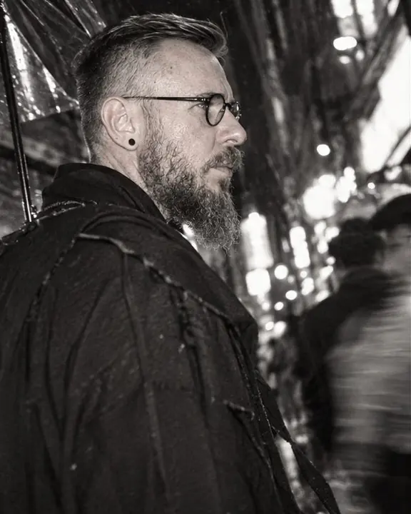 Profile of a bearded man in round glasses in a narrow Shinjuku alley, shop lights glowing as white blobs behind
