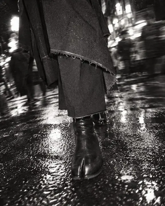 Low angle of a raw-hem black coat and split-toe boots on wet asphalt with reflections, grainy black and white