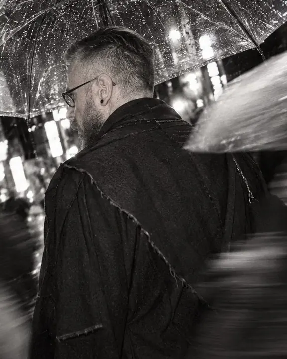 Rain beading on a clear umbrella next to the raw hem of a black wrap coat, high-contrast black and white