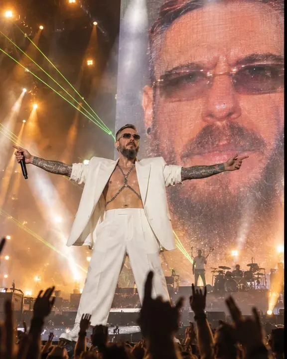 Wide view from the pit of a bearded man at the stage edge, arms spread, beams above and his face on a giant LED screen