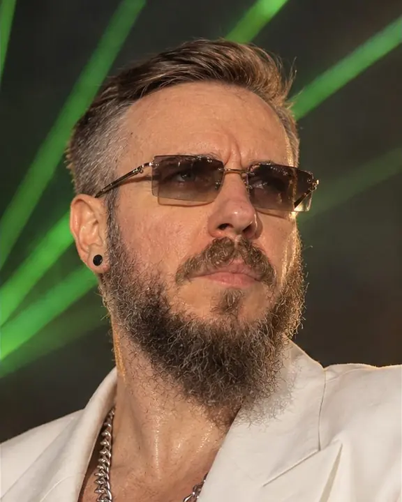 Telephoto close-up of a bearded man in a warm followspot, sweat shine on his face, green laser lines behind