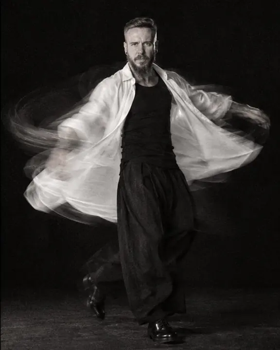 Black-and-white full-length of a bearded man spinning, his white shirt flaring into a curved trail, face frontal