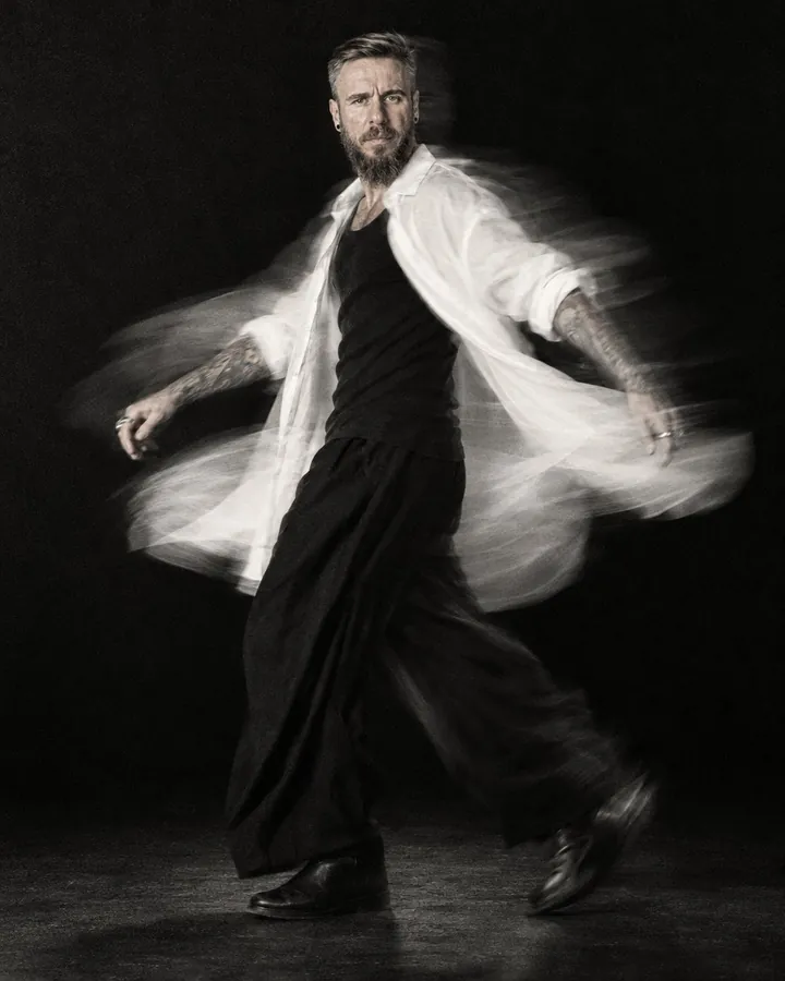 Black-and-white full-length of a bearded man spinning, his white shirt flaring into a curved trail, face frontal
