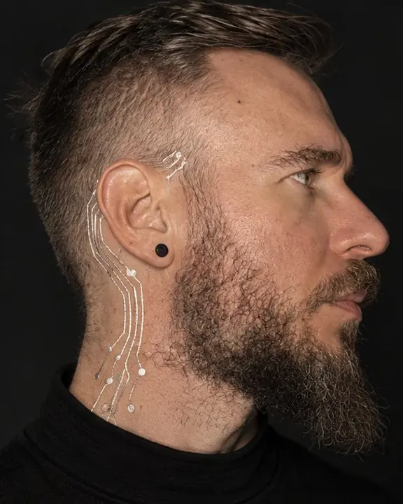 Tight profile of a bearded man, silver circuitry flowing from behind the ear down the neck in raking side light