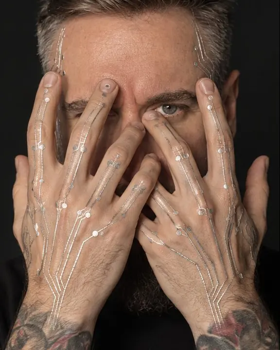 A bearded man raises his hands before his face, silver traces across the knuckles, eyes between his fingers