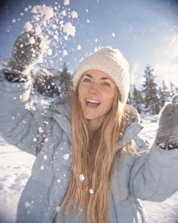 A woman with long blond hair laughs at the camera, mittened arms raised and snow flying, her ice-blue parka lit by flash