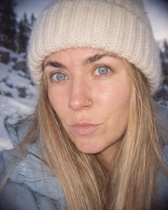 Off-center flash close-up of a woman with long blond hair under a cream beanie, eyes to the lens, the corners soft