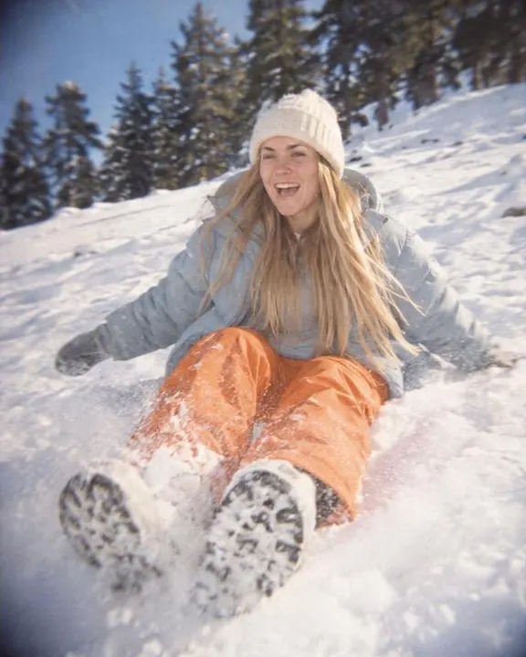 A woman with long blond hair slides down a gentle snowy slope in tangerine snow trousers, laughing, pines soft at the edge