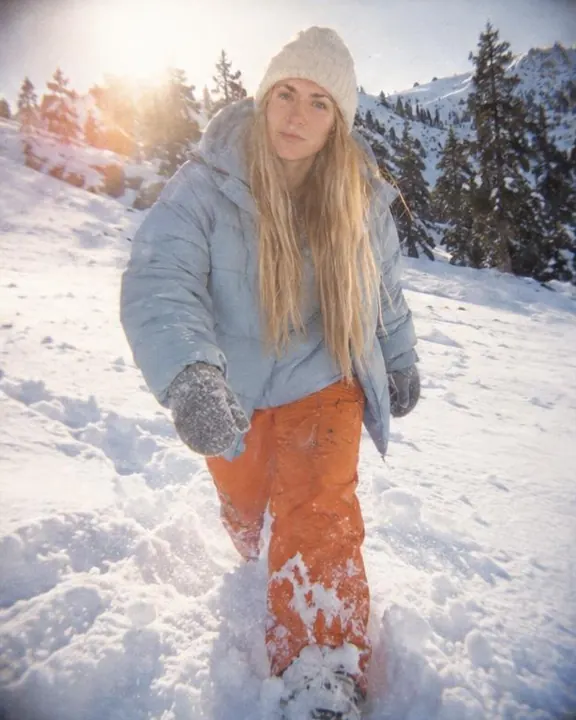 A woman with long blond hair wades through deep snow toward the camera, low sun behind her flaring orange in a corner