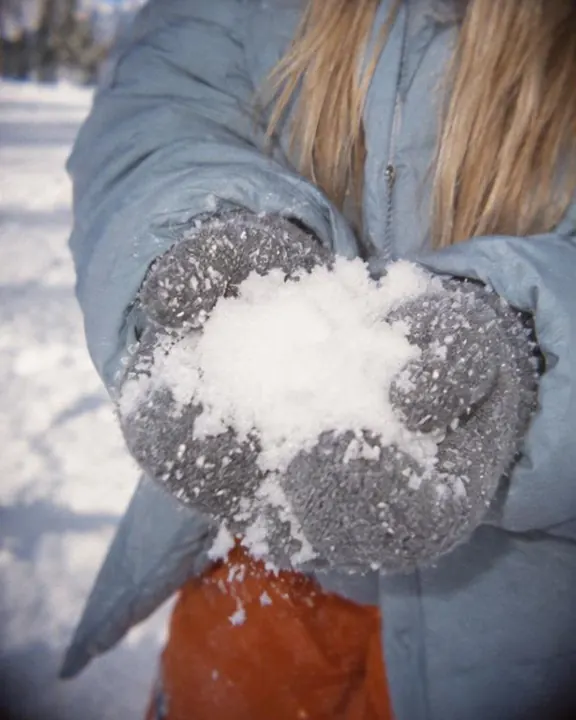A gray fleece mitten holding a handful of snow against a quilted ice-blue parka sleeve, flash sheen on the fabric