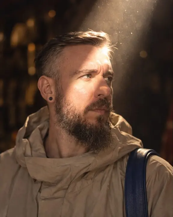 A bearded man's face lit inside a single shaft of sun in a dark Marrakech market, all else in shadow