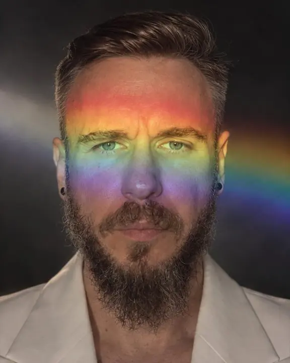 Head and shoulders of a bearded man with a rainbow band across his eyes, red above and violet at the chin