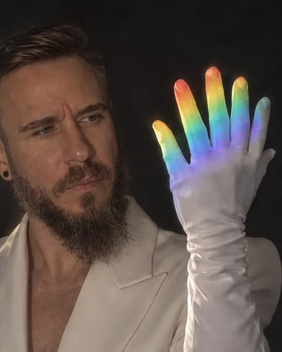 A man's raised hand in a white opera glove inside a beam of spectrum, colors falling across the fingers