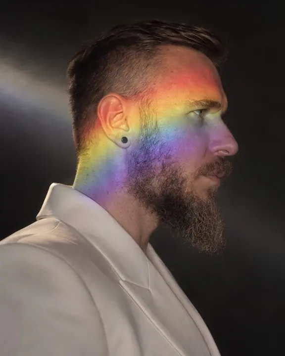 Profile of a bearded man in darkness, a rainbow band of projected light curving over his cheekbone